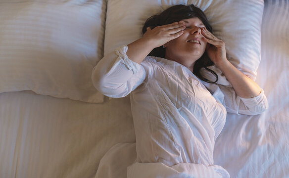 A woman with dark curly hair is sleeping in bed, wearing light-colored pajamas, on white striped bedding.