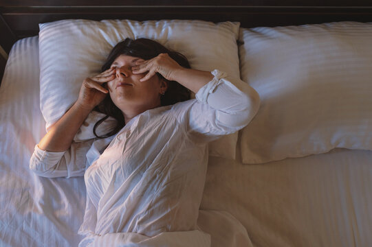 Waking up: a millennial woman sleeps in her bed at home, wearing light-colored pajamas, amidst white bedding, illuminated by the soft glow of a nightlight. A new day, a new morning.