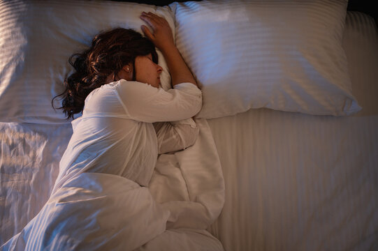 A millennial woman sleeps in her bed at home, wearing light-colored pajamas, under the glow of a nightlight on white bedding.