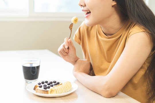 Happy young Asian woman smiling while enjoying a healthy breakfast with banana and blueberry toast with cream cheese, served with hot black coffee on a wooden table in the morning at home.