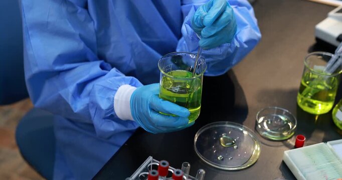 Laboratory worker in protective gown and gloves stirs bright green liquid in a glass beaker during a science experiment.