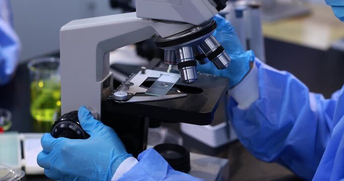 Close-up of a scientist wearing blue gloves and protective clothing adjusting a microscope in a laboratory, with a sample slide ready for examination.