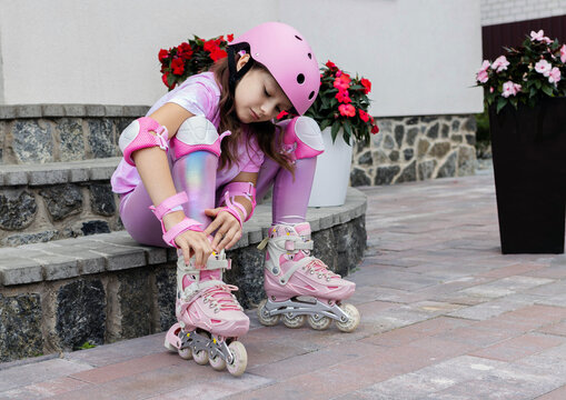 Close-up of a little girl in a pink helmet and protective gear fastening straps on her inline roller skates while sitting on stone steps outdoors. Rollerblading and active kid leisure concept