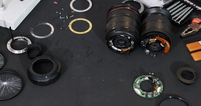 Top view of disassembled camera lenses, lens mounts, electronic parts, tools, and brushes on a dark workbench during optical equipment repair.