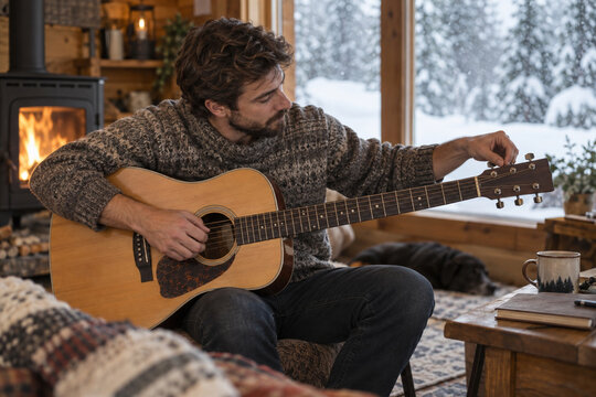 Adult tuning an acoustic guitar in a warm cabin with snowfall