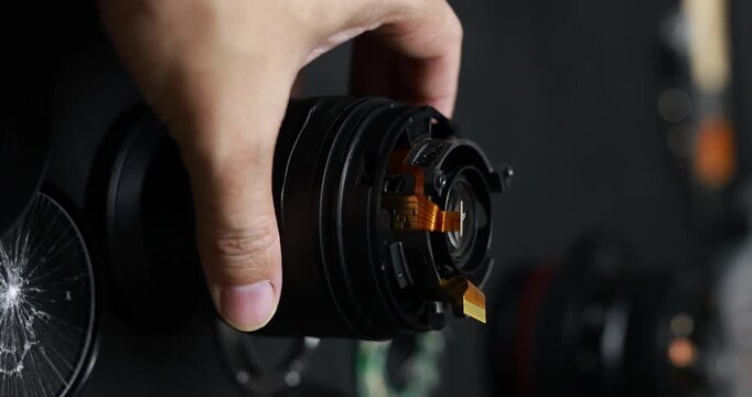 Close-up of a hand holding a disassembled camera lens with exposed electronic parts, repair and maintenance work in a dark workshop.