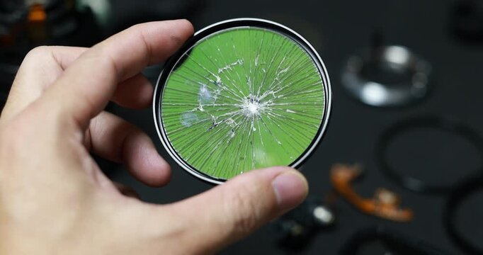 Close-up of a hand holding a cracked circular camera filter with shattered glass over a green background, damage, repair needs, and fragile equipment.