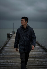 Fototapeta premium Loneliness and mental health, pensive man walking on a wooden pier during a stormy day with overcast sky and moody atmosphere for emotional reflection