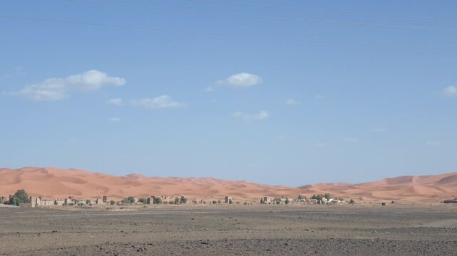 Driving past merzouga village with erg chebbi sand dunes in the background, experiencing the stunning and arid landscape of the moroccan sahara desert on a sunny day