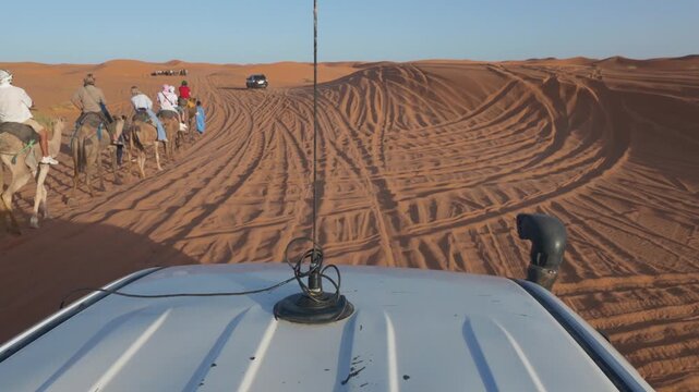Point of view of a four wheel drive vehicle dune bashing and driving on the sand dunes during a desert safari with a caravan of tourists riding camels in the background