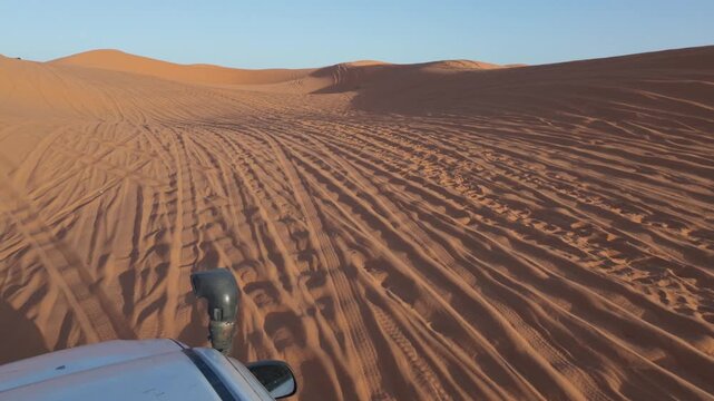 First person view of an off-road 4x4 vehicle driving through the vast arabian desert, leaving tire tracks on the golden sand dunes under a clear blue sky