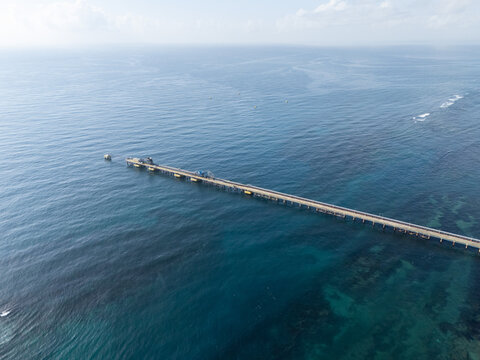 Aerial view of a long industrial pier extending into the deep blue Caribbean Sea, with clear water visible beneath the surface in Bajos de Haina, San Cristobal Province, Dominican Republic.