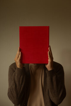 Front view portrait of a person holding a large red blank hardcover book in front of face, conceptual identity, storytelling and literature reader background on neutral beige studio wall.