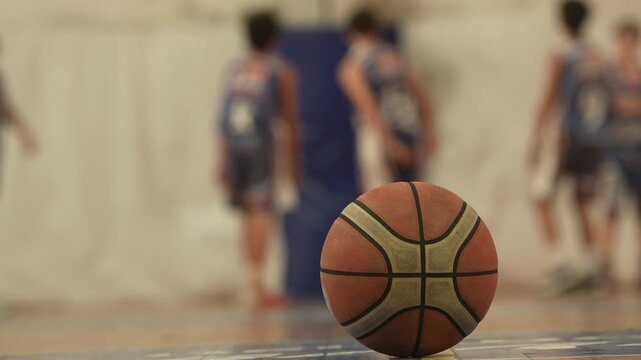 Basketball on indoor court floor in sharp focus with blurred unrecognizable players in background