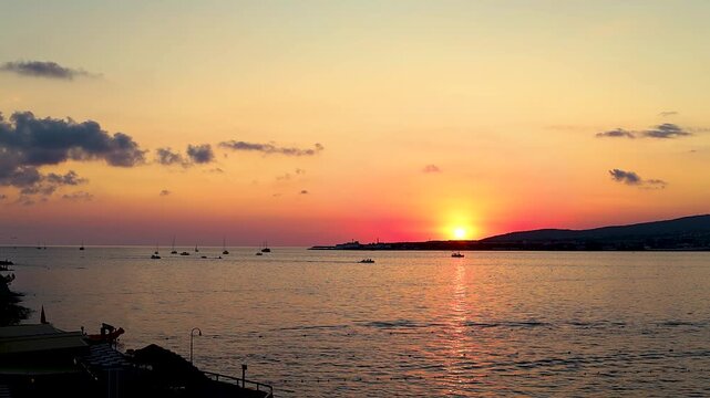 Sunset over calm water with boats sailing, silhouetted trees framing the scene, and distant hills creating a serene landscape in a coastal region