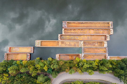 Aerial view of several rusted cargo barges moored along a riverbank lined with lush green trees and a winding road in Berlin, Berlin, Germany.