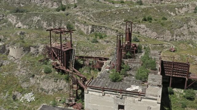 Aerial view of abandoned industrial factory with rusted metal structures and machinery set against the rugged terrain of the Caucasus Mountains, Russia.