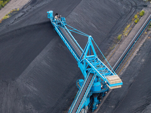 Aerial view of a large blue industrial stacker machine operating at a coal mining site with massive black coal piles and conveyor belt systems in Berlin, Berlin, Germany.