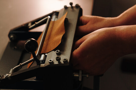 Artisan feeds leather through a manual skiving machine to reduce its thickness. Specialized craft process for premium accessories and handmade production.