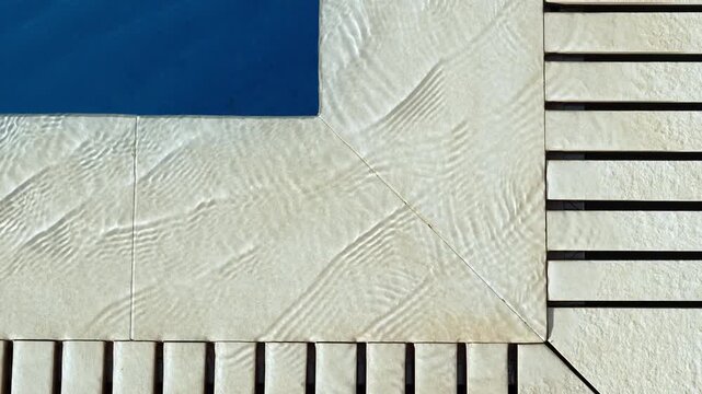 Top view of swimming pool edge with clear blue water rippling over stone tiles and wooden deck