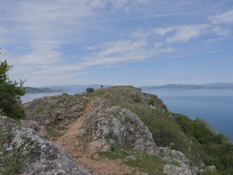 View of the rocky peninsula of Lin extending into Lake Ohrid, with a dirt path, sparse vegetation, and distant mountains under a blue sky in Lin, Korce County, Albania.
