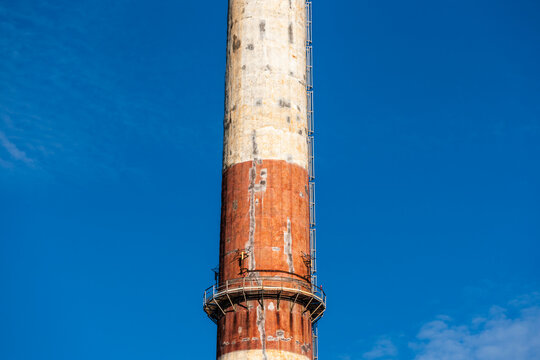 View of a tall industrial chimney with red and white painted concrete, a vertical ladder, and a metal maintenance platform against a clear blue sky in Poland.