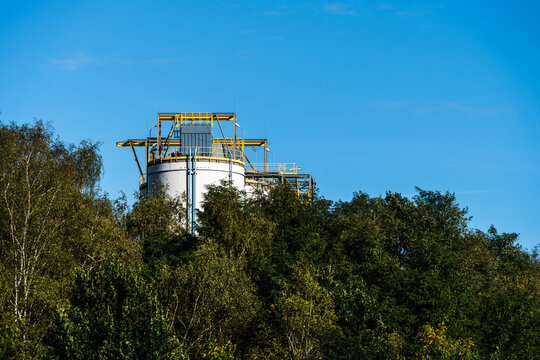 View of an industrial storage tank with a yellow metal framework on top, surrounded by dense green trees under a clear blue sky in Poland.