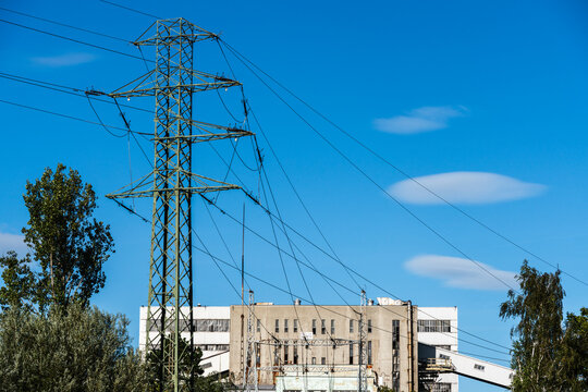 View of a green metal electricity transmission tower with power lines against a clear blue sky and an industrial building in the background in Poland.