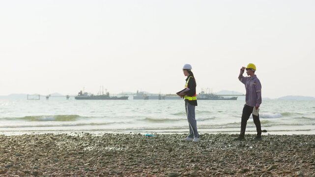 Two engineers conduct a site survey on a rocky shoreline, planning development for the offshore industrial port and energy facility in the background.