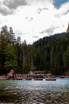 View of the wooden boathouse and rowing boats on the calm waters of Lago di Braies in the Dolomites, surrounded by dense pine forests under a cloudy sky in Rocca Pietore, Veneto, Italy.