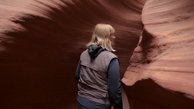 Rear view of blonde woman walking inside narrow passage of slot antelope canyon, admiring smooth spectacular and colorful sandstone curved rock formations carved by water erosion. Miracle of nature