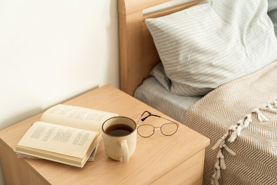 Cup of warm tea in mug on light wooden nightstand, an open book and reading glasses create cozy atmosphere for relaxation. Slow living, mindful hobby
