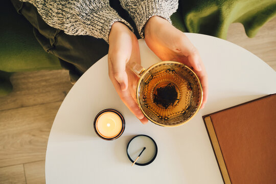 Person enjoying warm cup of tea with lit candle and book on side table