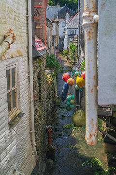 View of a narrow stream flowing between historic stone cottages, featuring colorful fishing buoys hanging near the water in Polperro, Cornwall, United Kingdom.