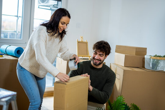 Happy young couple taping cardboard box together while moving into new home