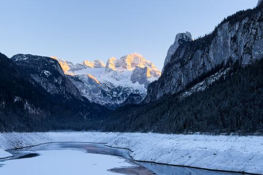 Cinematic sunset over Alpine lake in winter mountains. Winter mountain landscape. Golden hour reflection in calm mountain lake. Alpine peaks illuminated by orange light of setting sun.