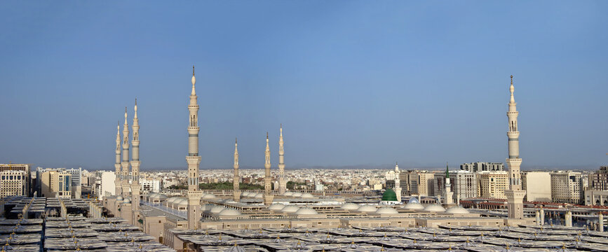 View of Al Masjid-e-Nabawi with its iconic minarets, green dome, and extensive courtyard umbrellas under a clear blue sky in Madinah, Al Madinah Province, Saudi Arabia.