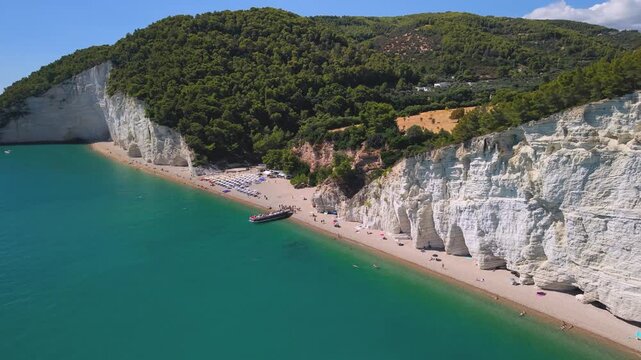 Vignanotica best beaches of Gargano Puglia Italy. aerial drone flyby white cliff organized beach umbrellas and tourist boat in turquoise sea. summer travel 4k video