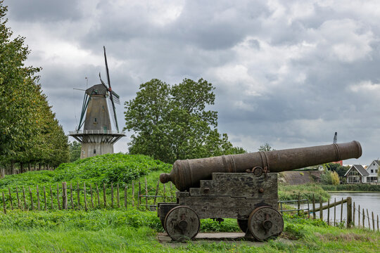 Woudrichem, Netherlands. Historic wooden cannon on ancient city fortress ramparts with traditional Dutch windmill under cloudy sky.