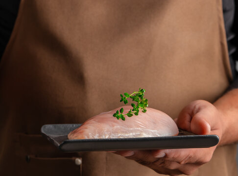 Raw chicken breast garnished with fresh herbs on a dark plate held by person wearing brown apron in close up culinary food preparation scene
