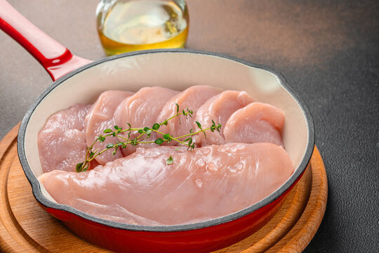 Raw chicken breast fillets arranged in a red enameled skillet with fresh thyme and olive oil on a wooden trivet, top view food ingredient