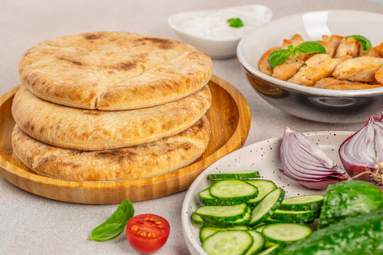 Stacked round flatbreads on wooden board with grilled chicken, yogurt sauce, cucumber slices, tomatoes, onions, and leafy greens in a tabletop food scene