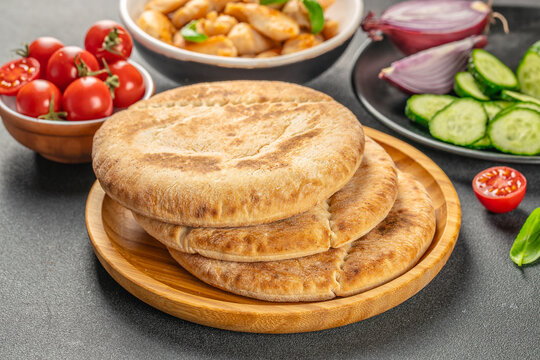Stack of round golden flatbreads on wooden serving board with tomatoes, cucumber, onion, greens, and cooked pieces in background, overhead food composition