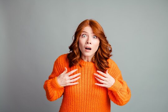 Surprised woman in orange sweater with shocked expression and hands to chest, red hair portrait in studio showing astonished emotional reaction and casual knit fashion