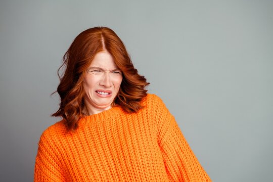 Woman in orange sweater making a disgusted face showing strong negative reaction and facial expression of dislike or surprise in a studio portrait with gray background
