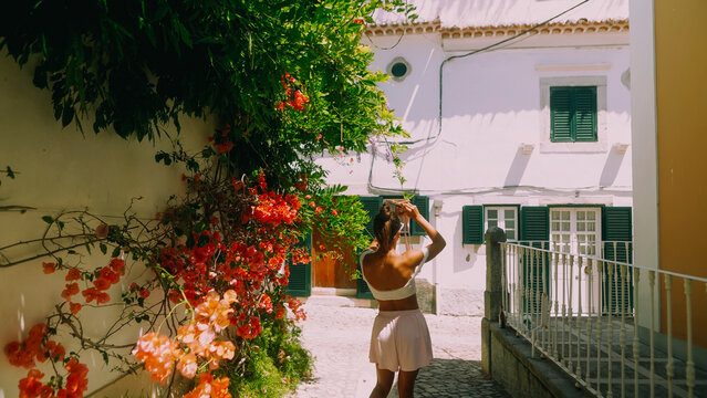 Young woman photographing sunlit Mediterranean alley with orange bougainvillea climbing beside white stucco buildings, green shutters, and cobblestone walkway