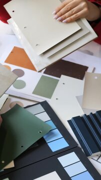 Hands review color swatches and sample boards around table, comparing stacked material sheets, finish cards, and texture samples for design selection during collaborative interior planning