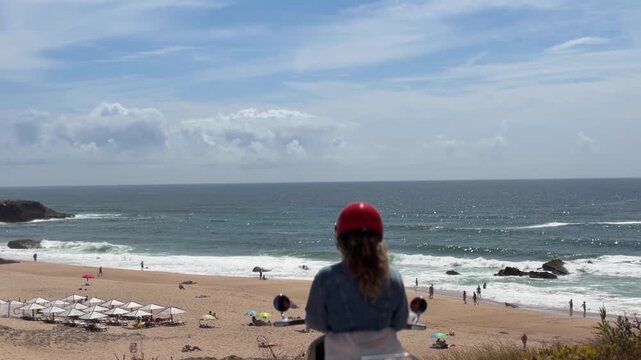 Rear view scooter rider with glossy red helmet and denim jacket looking toward sandy beach, foamy shoreline and ocean horizon under bright partly cloudy sky at coastal overlook