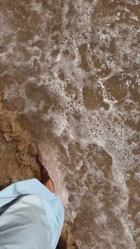 Overhead barefoot foot stepping into shallow foamy seawater on sunlit brown sand at beach shoreline with light blue shorts edge visible and sparkling wet sand highlights