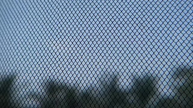 Smooth right-to-left panning video clip tracking a geometric wire mesh net or insect screen texture against a bright sky and blurry tree silhouettes. Ideal for abstract backgrounds, security, enclosur